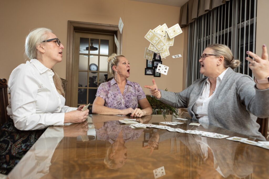 Three women sitting at a table playing cards. Cards tossed into the air.