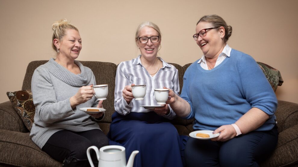 Three women seated on a sofa holding tea cups.